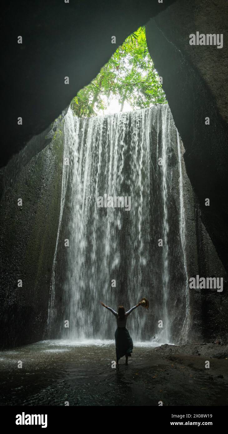 Eine glückselige Szene fängt eine Person mit erhobenen Armen vor einem atemberaubenden Wasserfall ein, umgeben von üppiger Vegetation im Herzen von Bali. Stockfoto