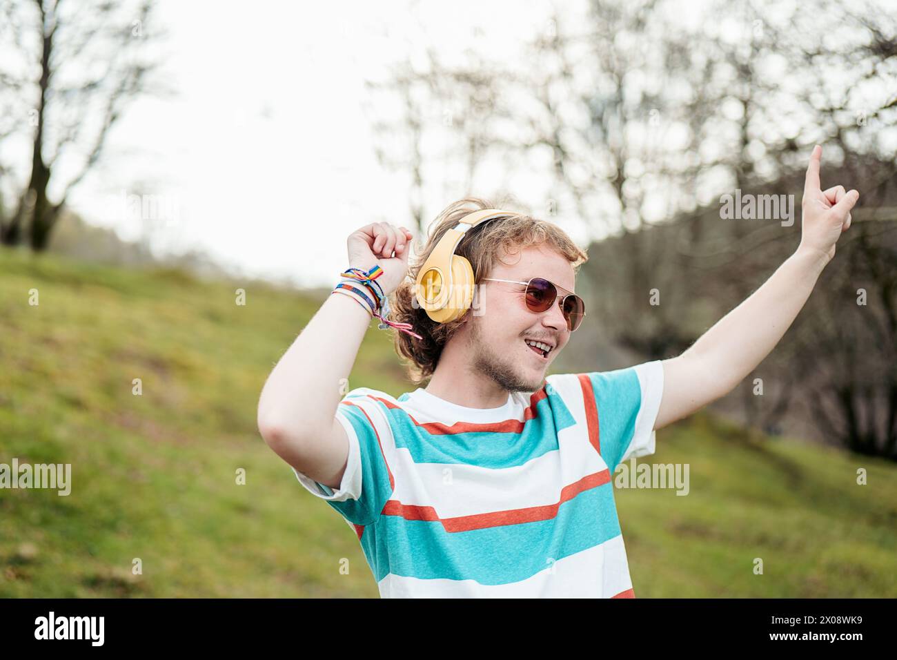 Ein fröhlicher Mann mit einem breiten Lächeln und gelben Kopfhörern hebt seine Hand vor Freude, während er in einer ruhigen Umgebung im Freien tanzt Stockfoto