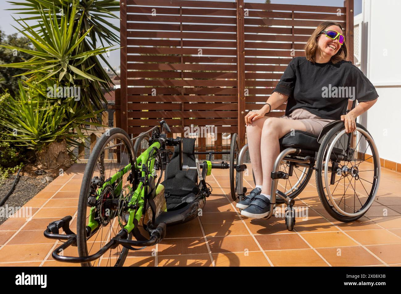 Eine lächelnde Frau in Sonnenbrille sitzt in ihrem Rollstuhl neben ihrem grünen Handrad und zeigt Entschlossenheit und Mobilität. Stockfoto