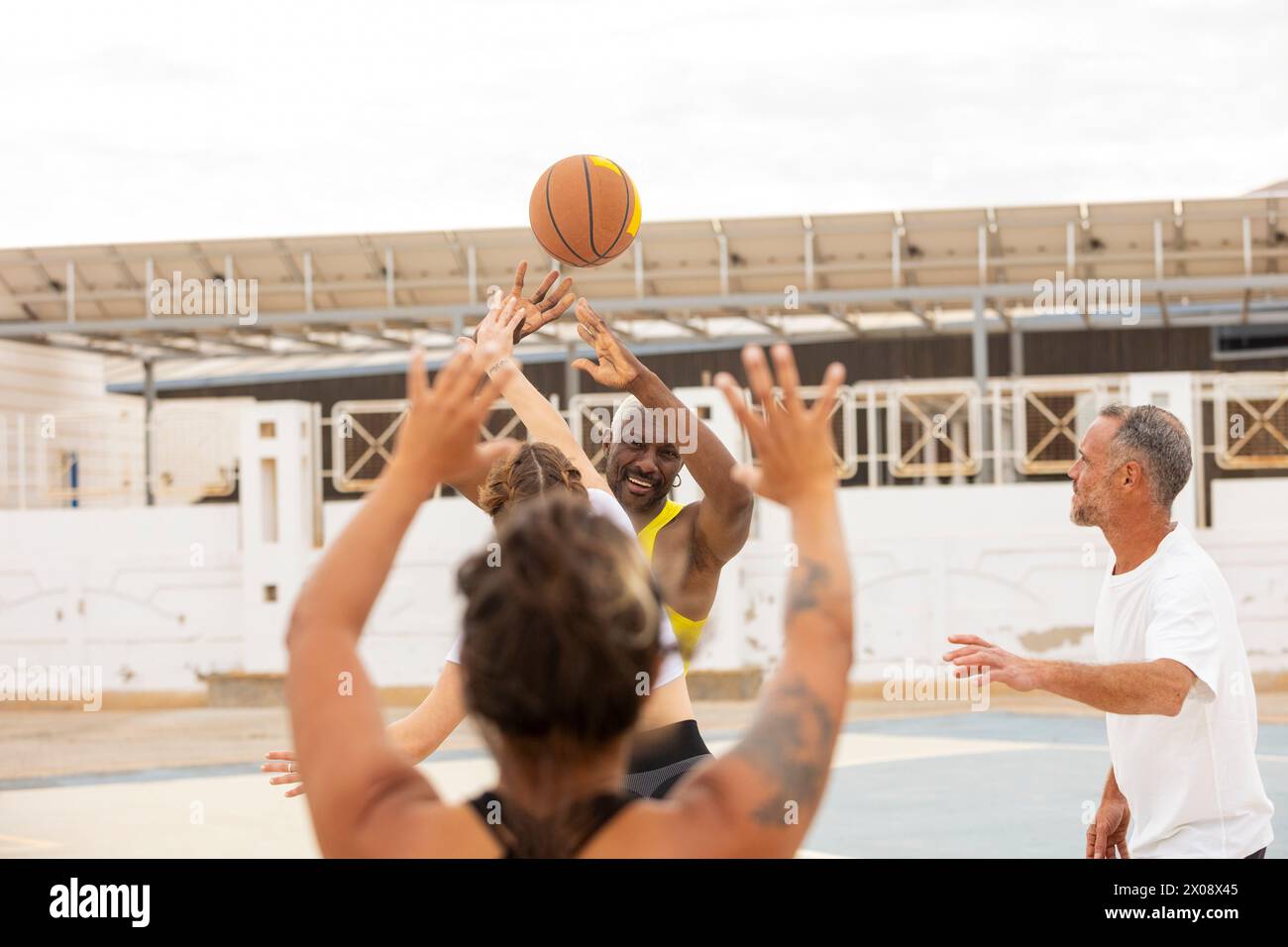 Ein Basketballspieler in einem gelben Tanktop macht einen Schuss, während seine Teamkollegen in einem dynamischen Street Basketballspiel eifrig nach draußen greifen und blockieren Stockfoto
