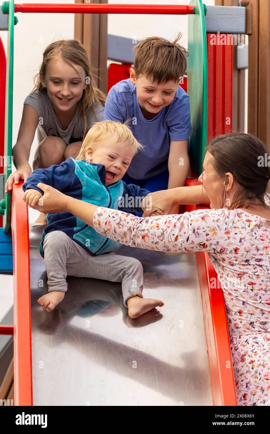 Eine lebendige und herzerwärmende Szene, während eine Familie die Zeit zusammen auf einer Spielplatzrutsche genießt. Die pflegende Mutter hilft dem Kind, das begeistert ist, und zeigt eine Funktion Stockfoto