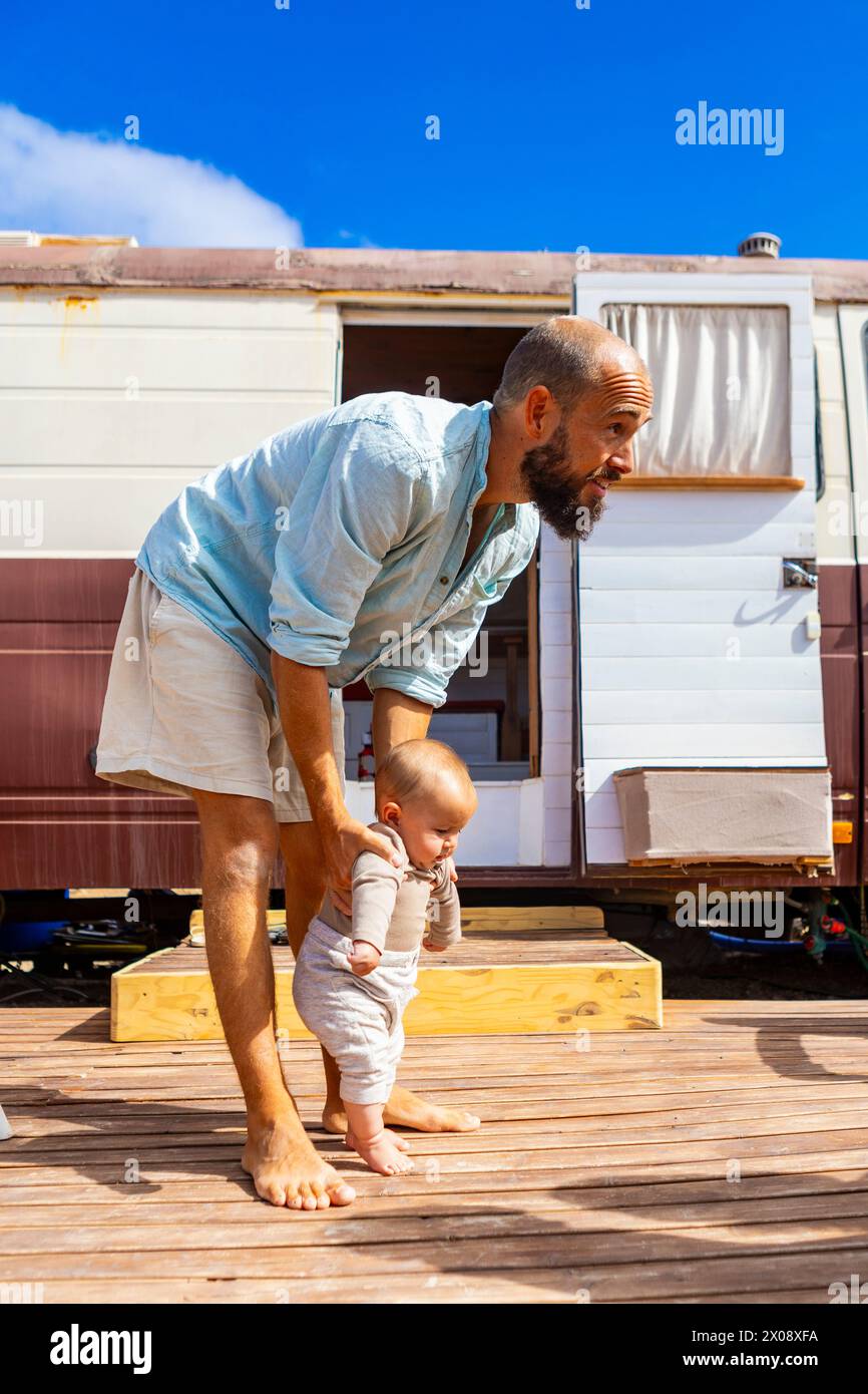 Der Mann hilft einem Baby, die ersten Schritte auf einer Holzterrasse vor dem Familienbus zu machen. Stockfoto