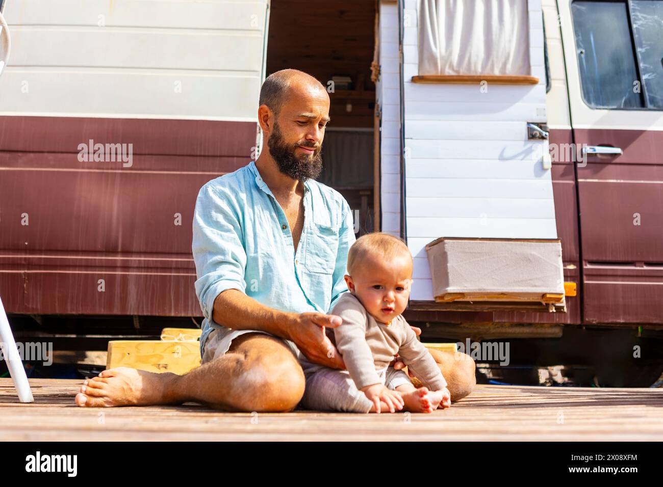 Ein Mann und ein Baby sitzen auf einer hölzernen Terrasse neben ihrem Haus auf Rädern und symbolisieren nachhaltiges Leben und Familienbindung. Stockfoto