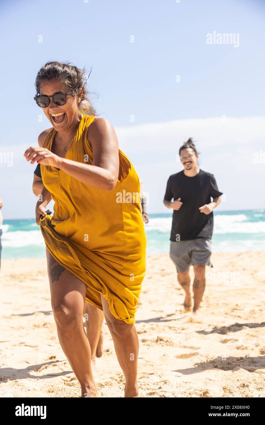 Eine multiethnische Frau in gelbem Kleid lacht fröhlich, während sie mit einem multiethnischen Mann im Hintergrund auf dem Sand läuft. Stockfoto