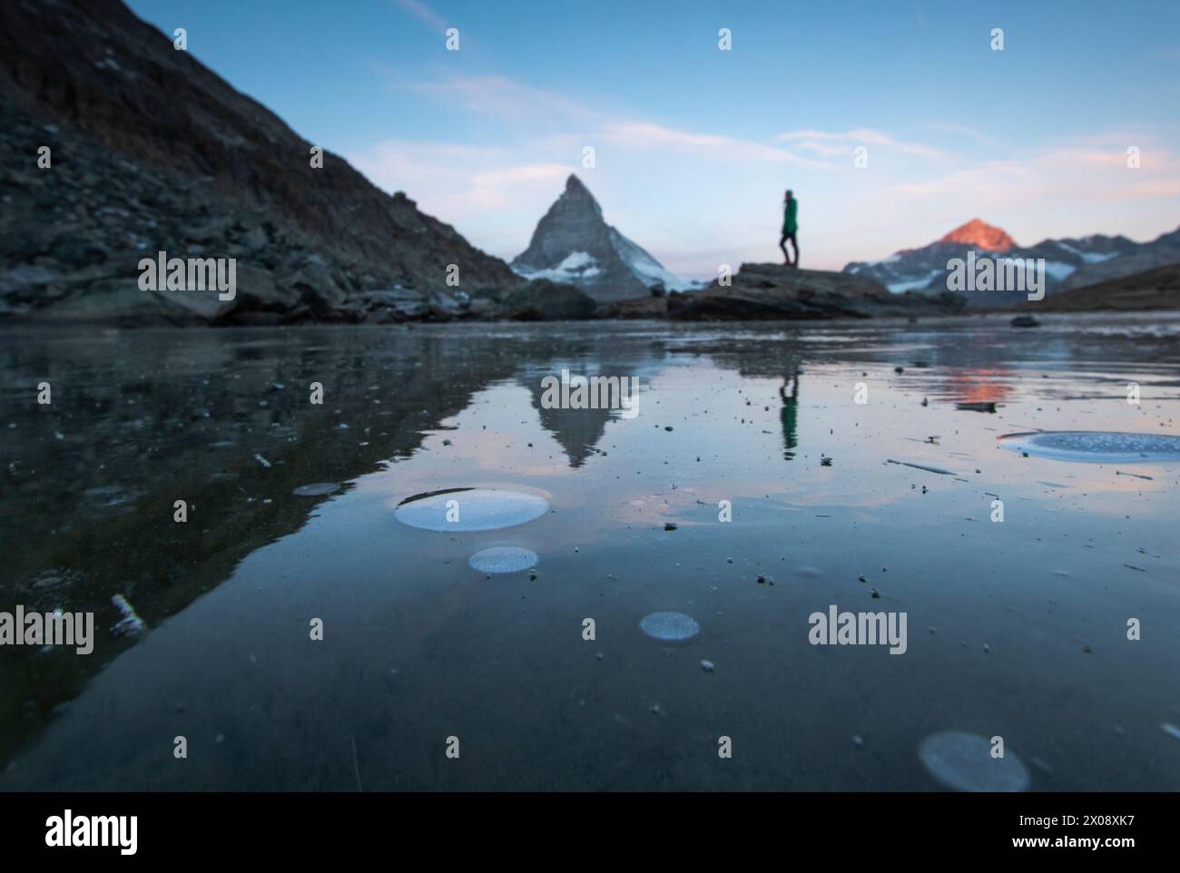 Eine ruhige Frühlingsszene fängt einen teilweise aufgetauten See ein, der das berühmte Matterhorn in den Schweizer Alpen reflektiert, mit einer nicht erkennbaren Person in s Stockfoto