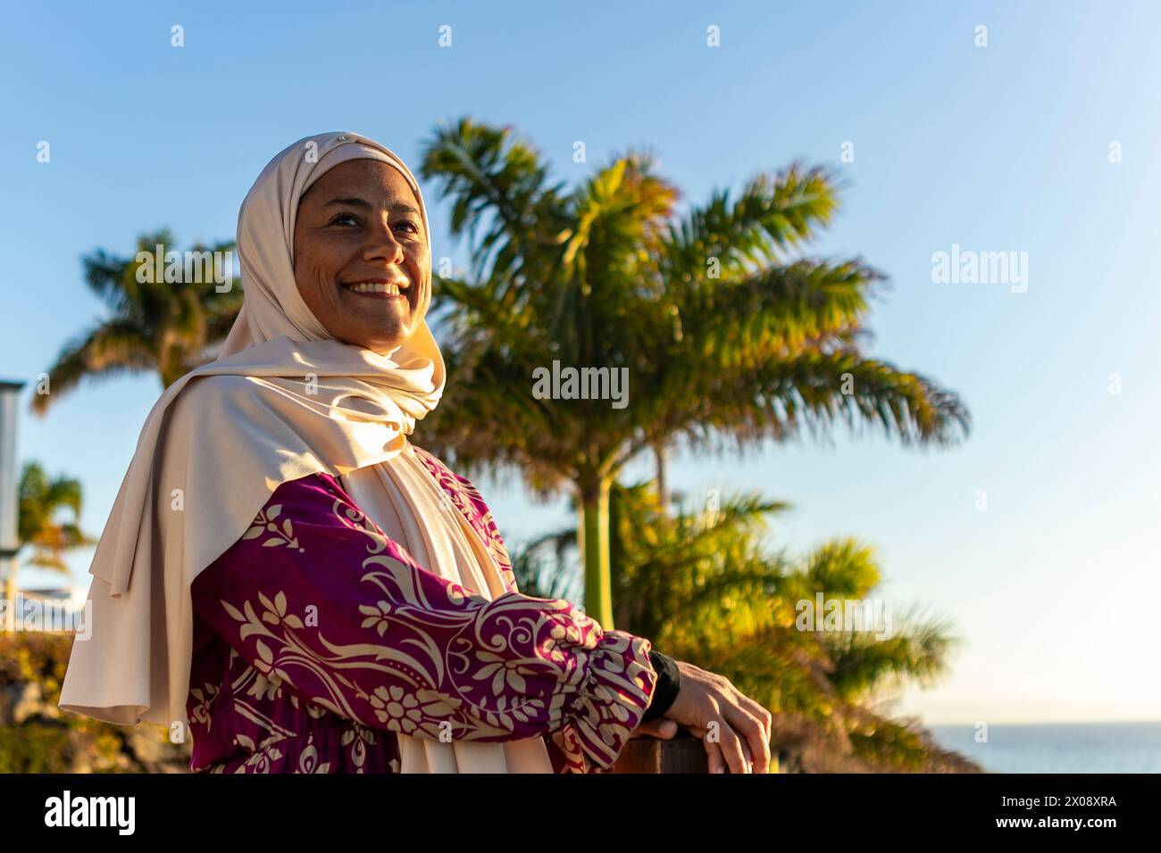 Eine fröhliche muslimische Frau, die einen weißen Hijab und ein gemustertes Kleid trägt, lächelt warm, während sie am Meer unter einem klaren blauen Himmel steht Stockfoto