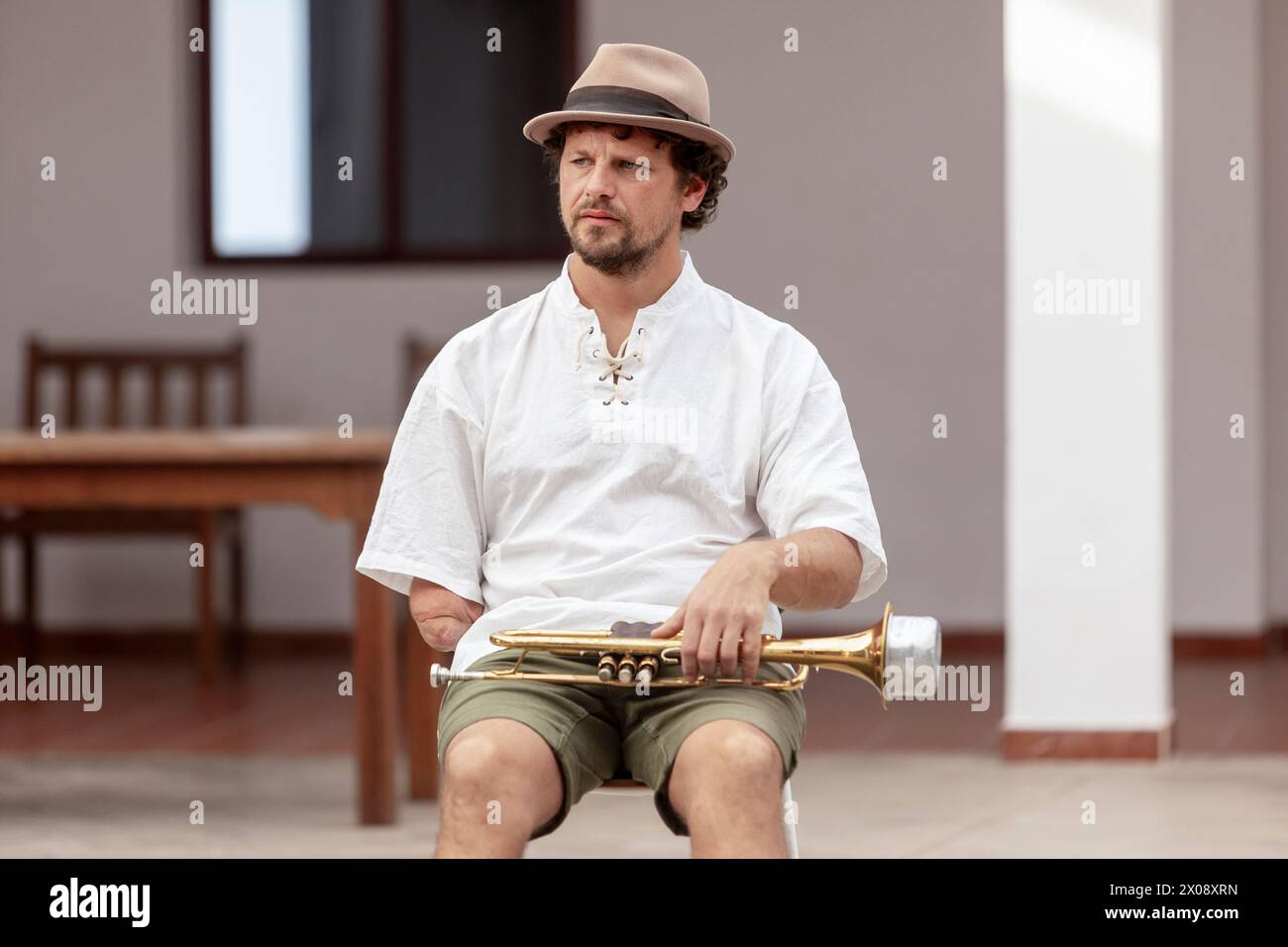 Ein Mann sitzt draußen auf einem Hocker, hält eine Trompete und trägt legere Kleidung, die ein entspanntes Ambiente mit musikalischem Thema weckt Stockfoto