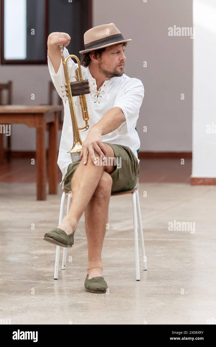 Ein einarmiger Mann mit Hut und lässiger Kleidung übt das Trompetenspiel, während er auf einem Stuhl sitzt Stockfoto