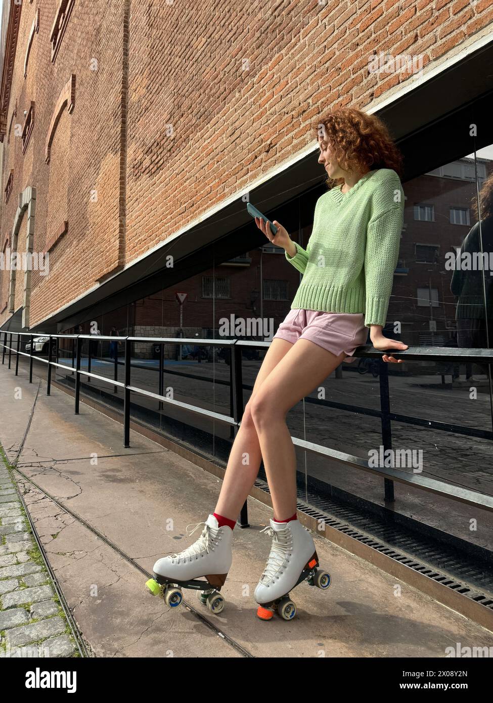 Eine junge rothaarige Frau in lässiger Kleidung Rollschuhlaufen, während sie mit ihrem Smartphone interagiert, ein Stadtbild mit Industriearchitektur, der das umrahmt Stockfoto