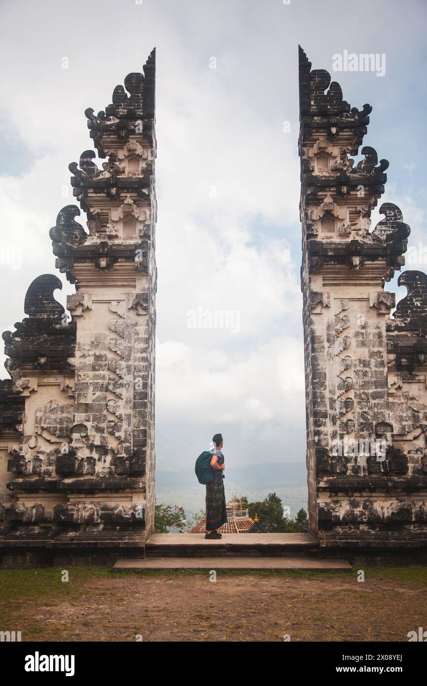 Ein Reisender steht zwischen den traditionellen geteilten Toren, Candi Bentar von Bali, symbolisiert den Eingang zum Heiligen, gekleidet in lokaler Kleidung und Cheri Stockfoto