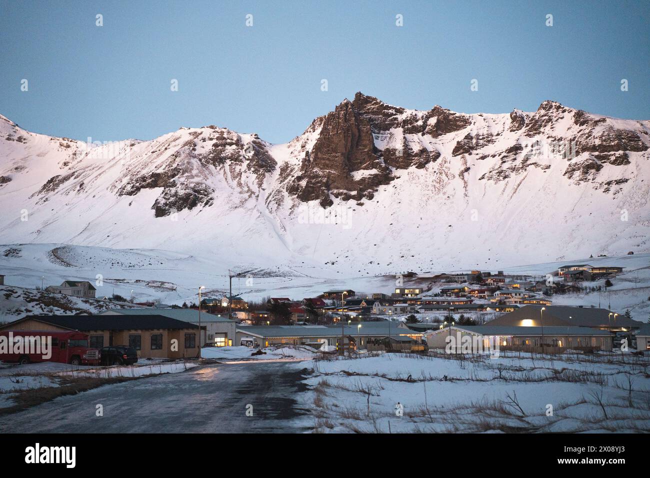 Ein gemütliches isländisches Dorf schmiegt sich am Fuße der hoch aufragenden, schneebedeckten Berge, beleuchtet von der sanften Abenddämmerung Stockfoto