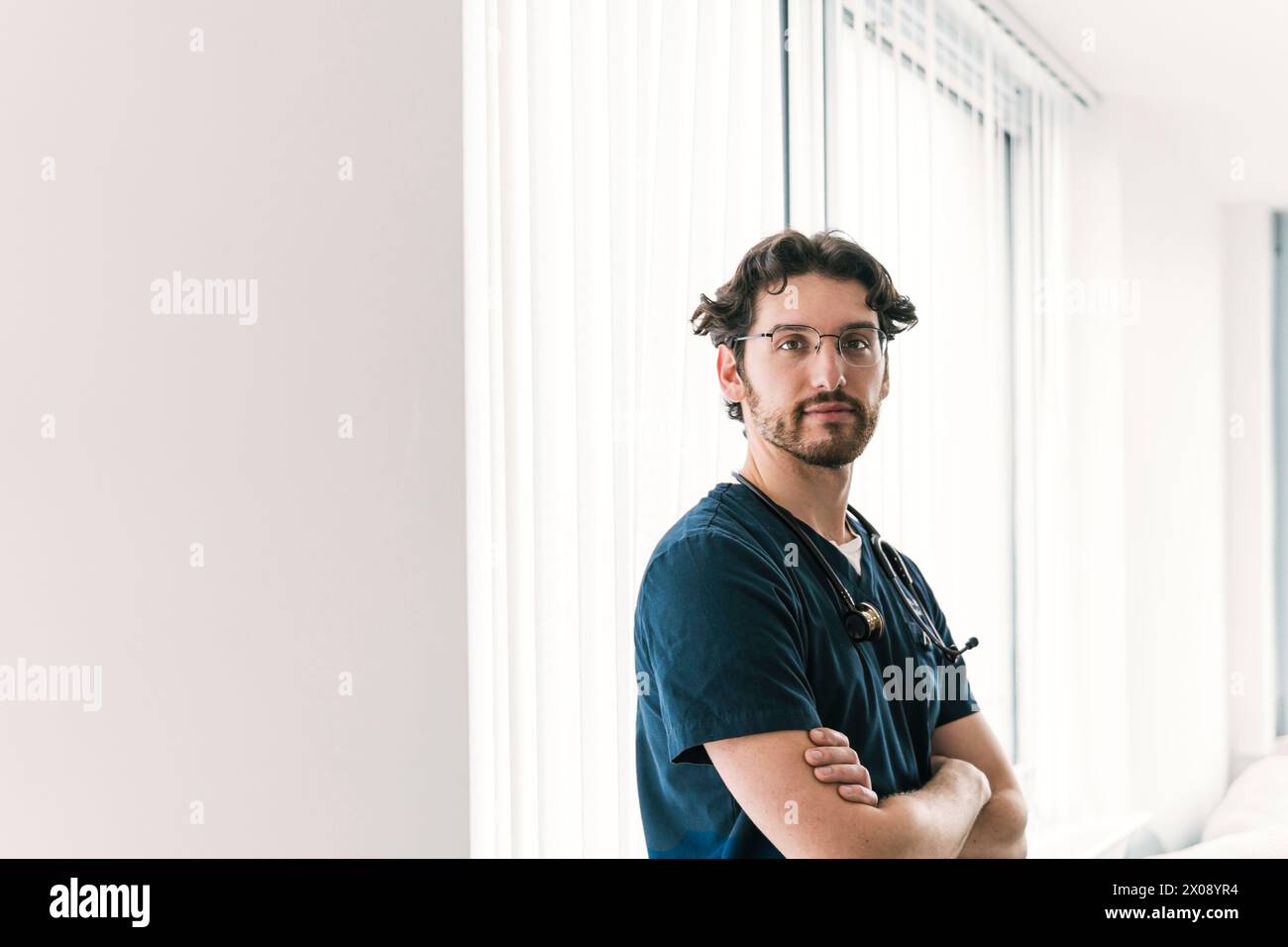 Ein ruhiger männlicher Mediziner steht mit gefalteten Armen in einem hellen, modernen Klinikumfeld und strahlt ruhige Kompetenz aus Stockfoto