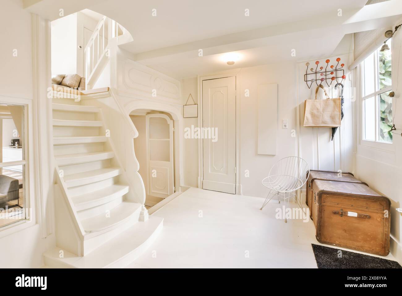 Ein gut beleuchteter, weiß gestalteter Eingangsbereich mit einer Wendeltreppe, einem Vintage-Koffer und dekorativen Wandhaken. Stockfoto