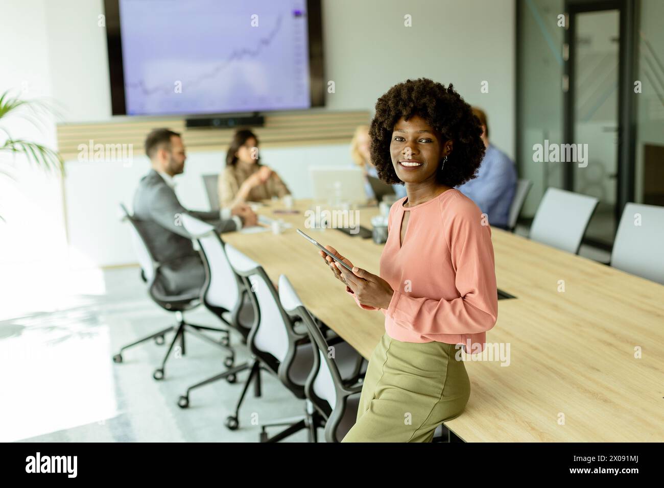 Ccheerful Afroamerikanerin leitet ein Meeting und hält sich an einem digitalen Tablet fest, während Kollegen hinter ihr Gespräche führen. Stockfoto