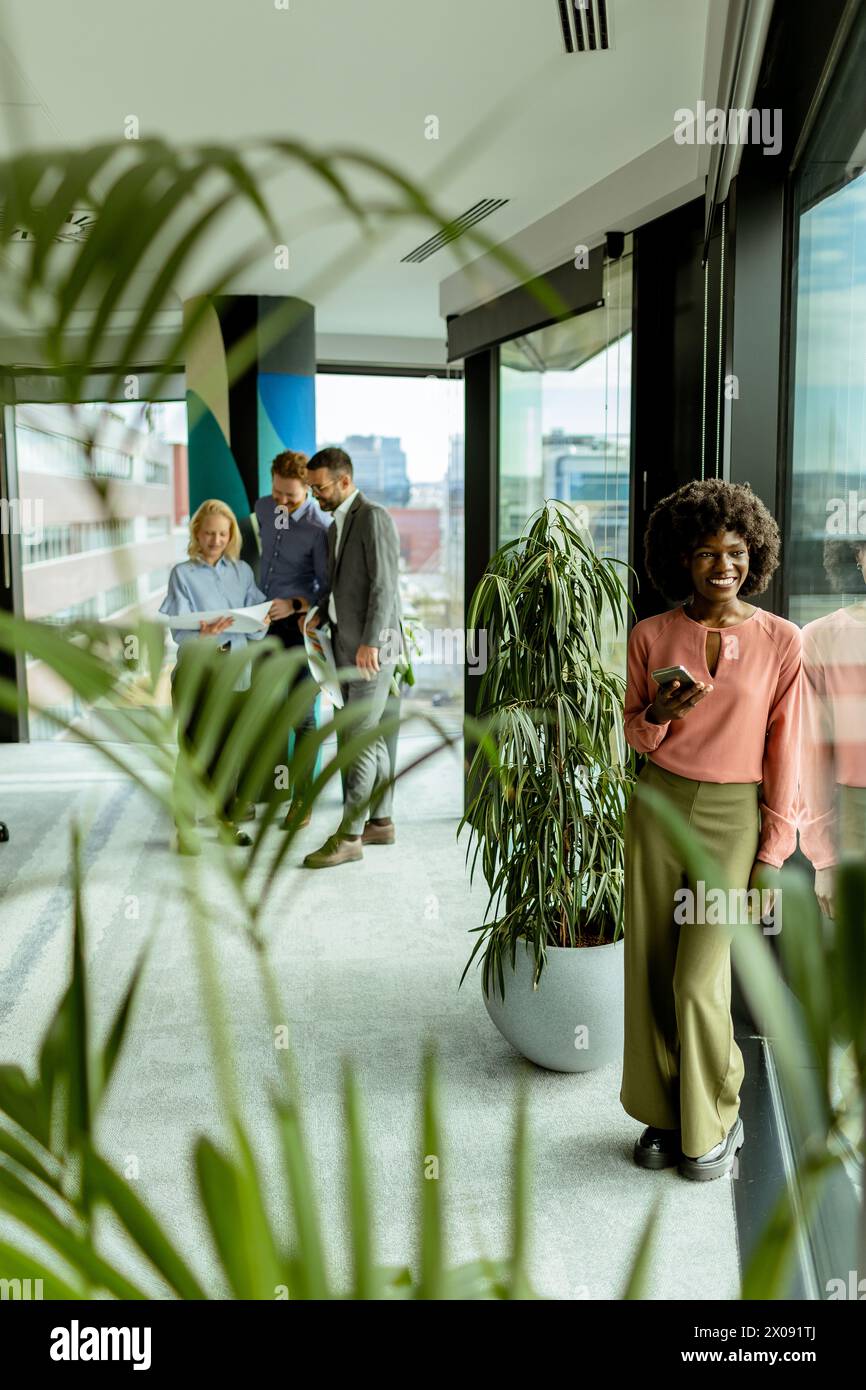 Die afroamerikanische Frau am Telefon steht neben üppigen Pflanzen im Haus, während sich die Kollegen im Hintergrund unterhalten Stockfoto