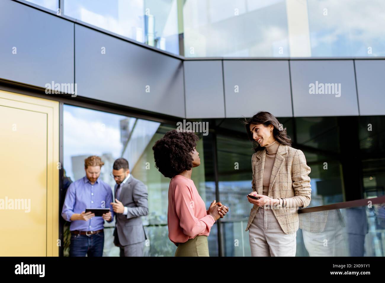 Zwei weibliche multiethnische Kollegen unterhalten sich vor einem Büro freundlich, während andere im Hintergrund arbeiten Stockfoto