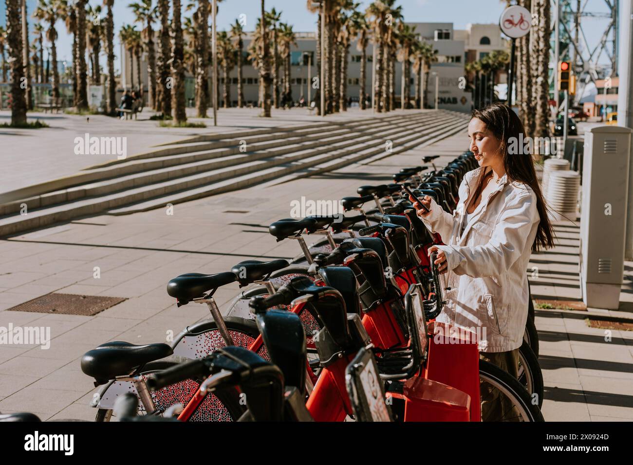 Die junge Frau steht bei einer Reihe roter Fahrräder im sonnigen Barcelona und überprüft ihr Handy, wahrscheinlich mit einer Bike-Sharing-App Stockfoto