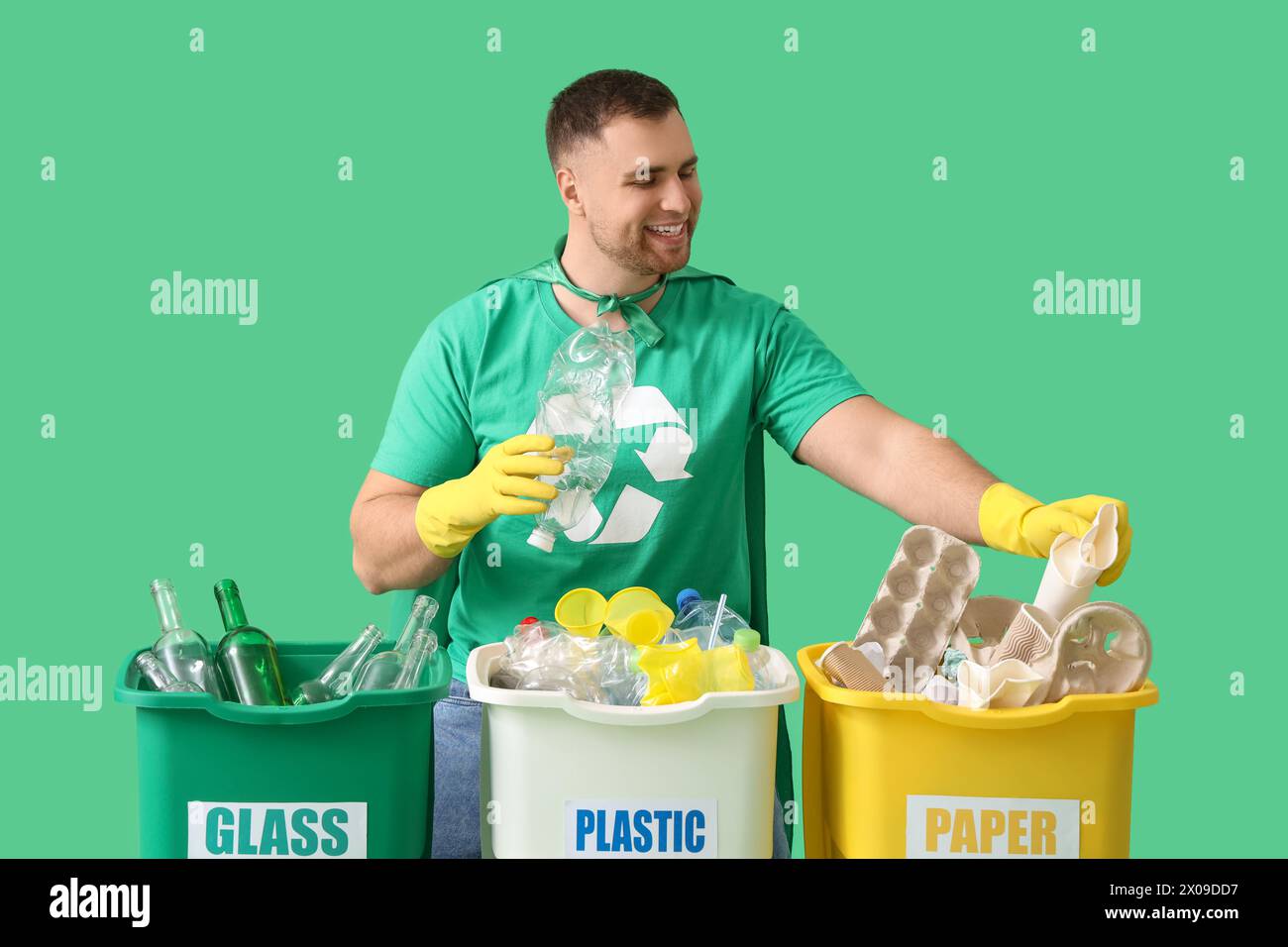 Männlicher Freiwilliger im superheldenumhang mit Papierkorb, die Müll auf grünem Hintergrund sortieren Stockfoto