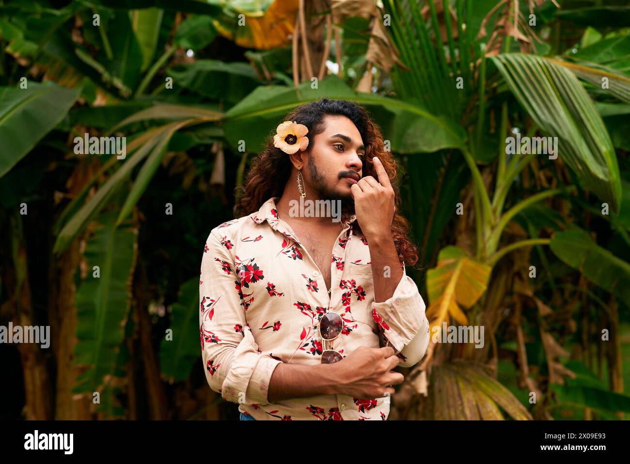 Sri-lankischer Mann mit Blume im Haar, steht im tropischen Garten und trägt ein Blumenhemd. Verkörpert Geschlechterfluidität, weiblichen Ausdruck, LGBT-Stolz Stockfoto