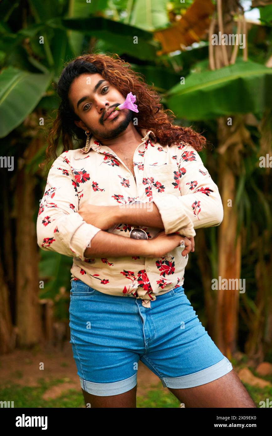 Selbstbewusster schwuler Mann mit lockigen Haaren, Blume im Mund, steht im tropischen Garten. Drückt queeren Stolz aus, Geschlechterfluidität. Shorts und Hemd mit Blumenmuster Stockfoto