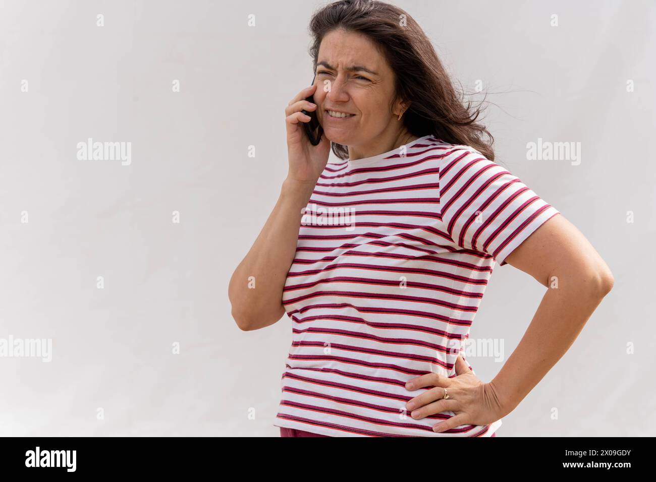 Weiße Frau mittleren Alters mit langen Haaren, die sich auf einem Handy unterhalten. Sie ist stilvoll in einem gestreiften Oberteil vor einem hellen Hintergrund gekleidet. Stockfoto