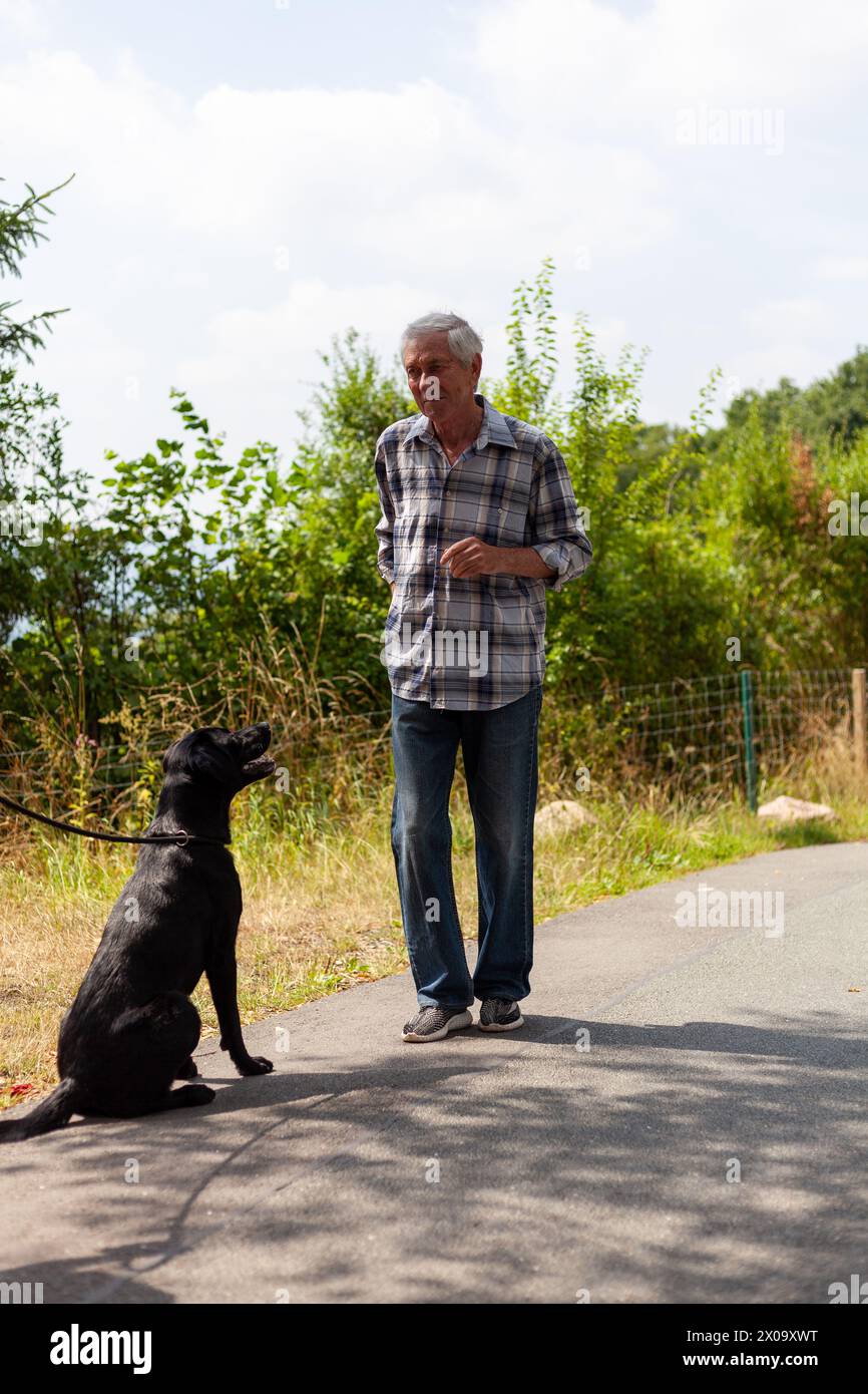 Besondere Verbindung zwischen einem älteren Mann und seinem schwarzen Labrador-Gefährten, die einen friedlichen Moment gemeinsam auf einer Landstraße genießen Stockfoto