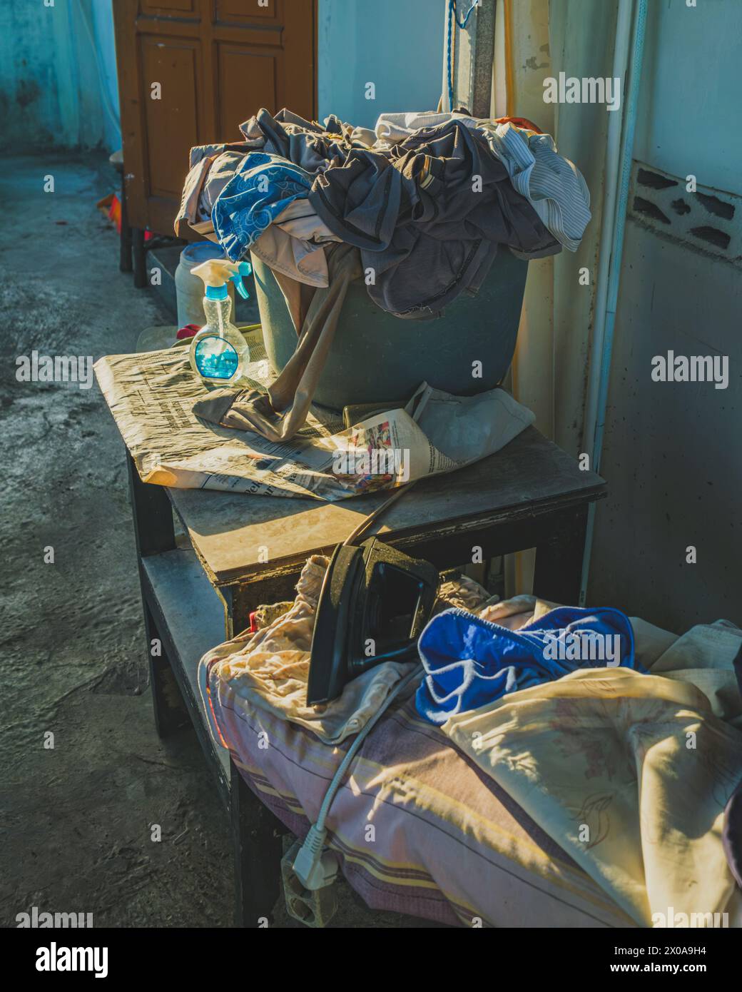 Auf einem hellbraunen Holztisch steht ein Wäschekorb mit bunten Kleidern. Ein blaues Bügeleisen und eine weiße Sprühflasche liegen rechts vom Ba Stockfoto
