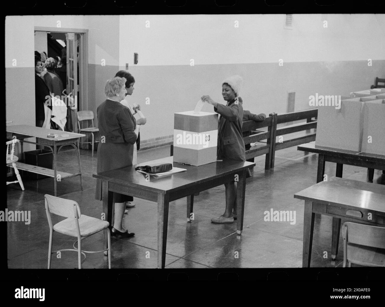 Das Foto zeigt eine junge Frau, die ihren Wahlzettel an der Cardozo High School abgibt, die von Wahlhelfern überwacht wird, während andere Wähler draußen in einem Flur warten, Washington, District of Columbia, 3. November 1964. Foto von Marion S Trikosko/US News and World Report Collection) Stockfoto
