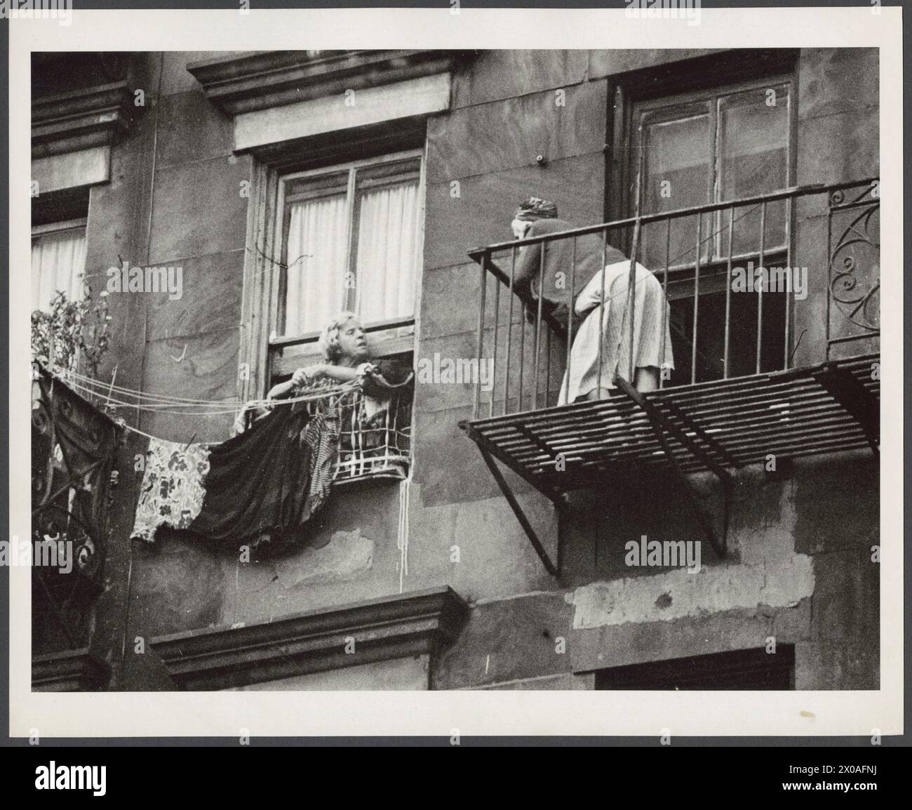 Zwei Frauen, die sich aus dem oberen Stockwerk eines Wohnhauses unterhalten; Frau links pinnt Wäsche in die Wäscheleine, eine rechts steht auf der Feuerkelwand, New York, New York, September 1955. (Foto: Anthony Rizzuto/Anthony Angel Collection) Stockfoto