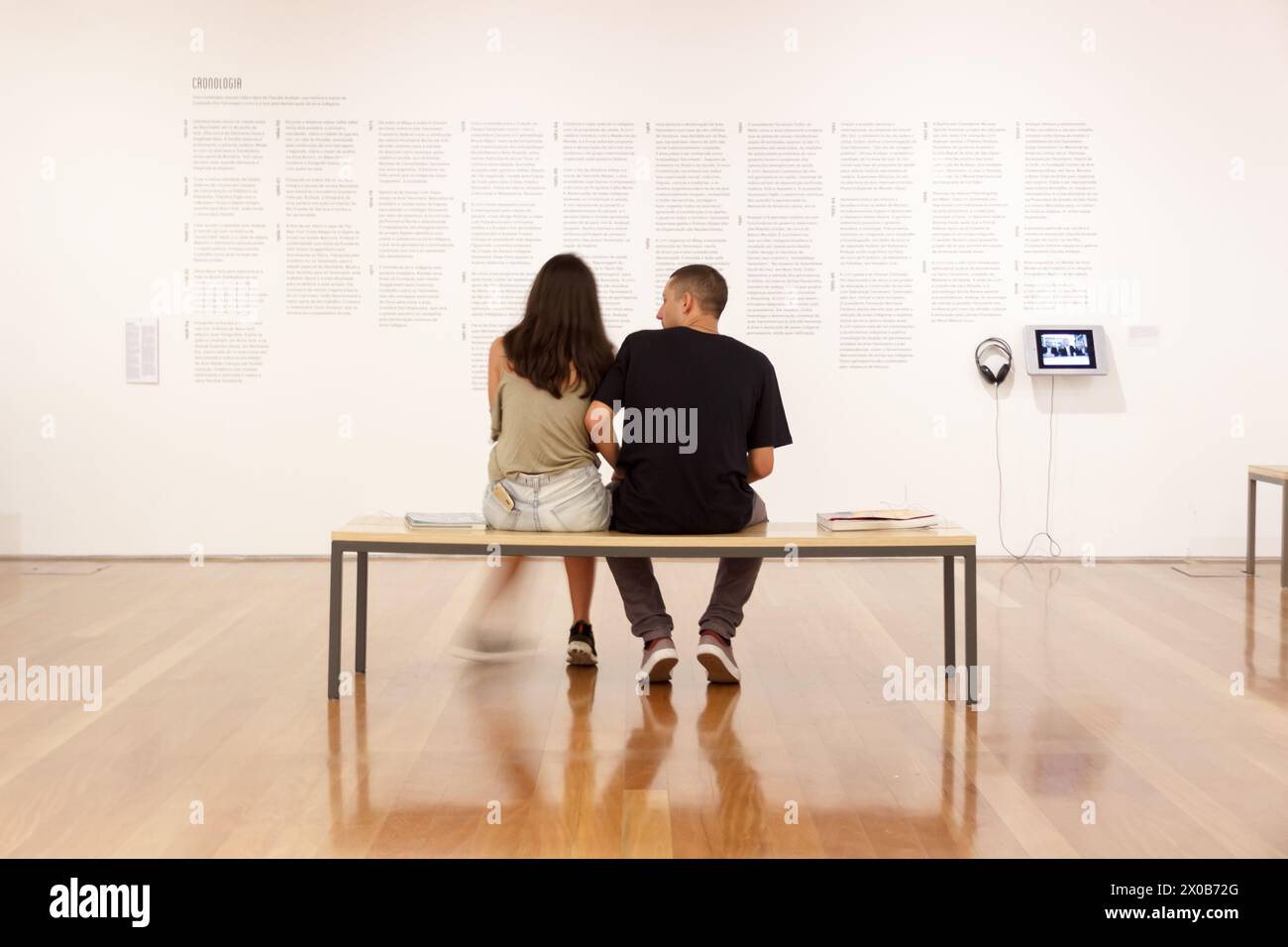 Ein Paar, das auf einer Bank in einer Museumsausstellung sitzt, lacht und auf die Wand mit Kunstwerken blickt Stockfoto