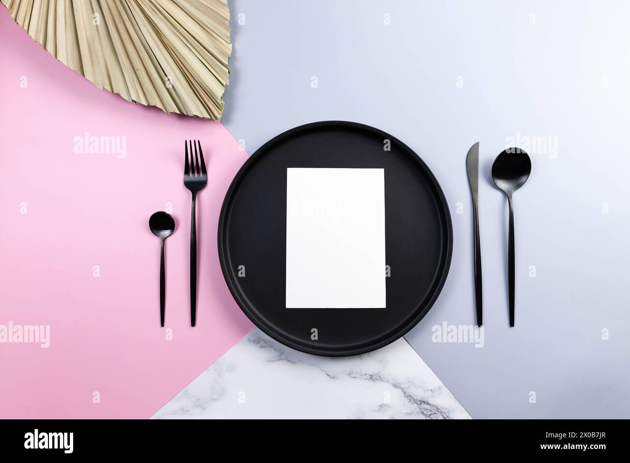 Draufsicht auf schwarzen Teller und Besteck, weiße Karte, getrocknete Palmblätter auf farbenfrohem Hintergrund. Tischdekoration, blau, rosa und Marmorhintergrund. Pastellfarbe Stockfoto
