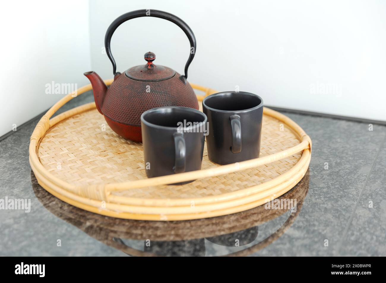 Detail einer roten chinesischen Teekanne und zwei schwarze Tassen auf einem Korbtablett in einer hellen Küche. Niemand drinnen Stockfoto