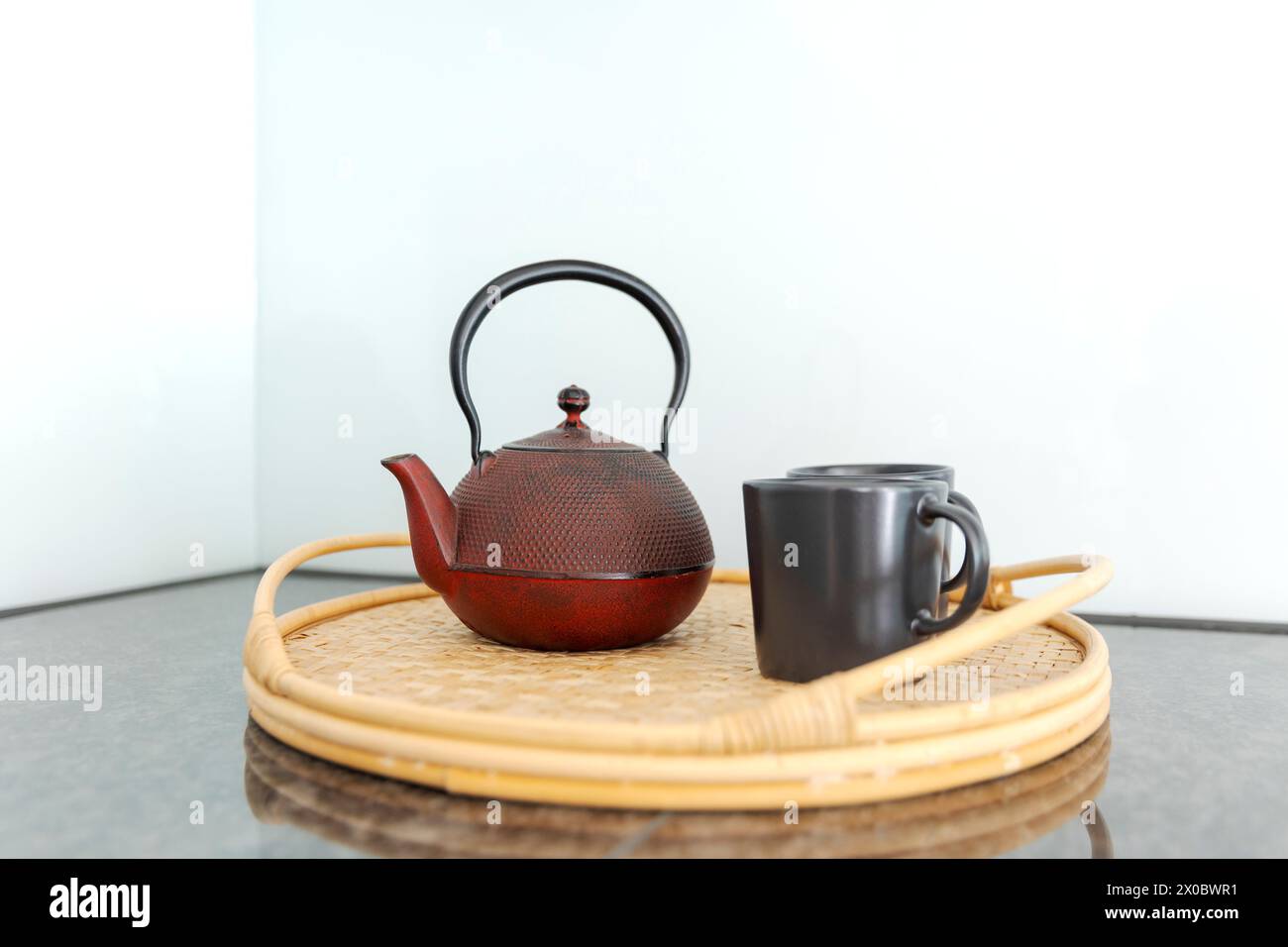 Detail einer roten chinesischen Teekanne und zwei schwarze Tassen auf einem Korbtablett in einer hellen Küche. Niemand drinnen Stockfoto