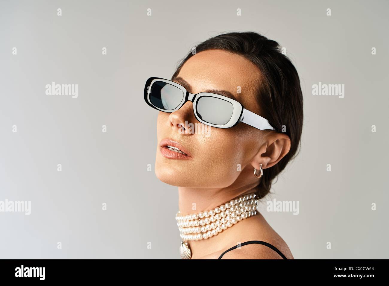 Eine stilvolle Frau mit Sonnenbrille und Perlenkette verströmt Vertrauen in ein Studio vor grauem Hintergrund. Stockfoto