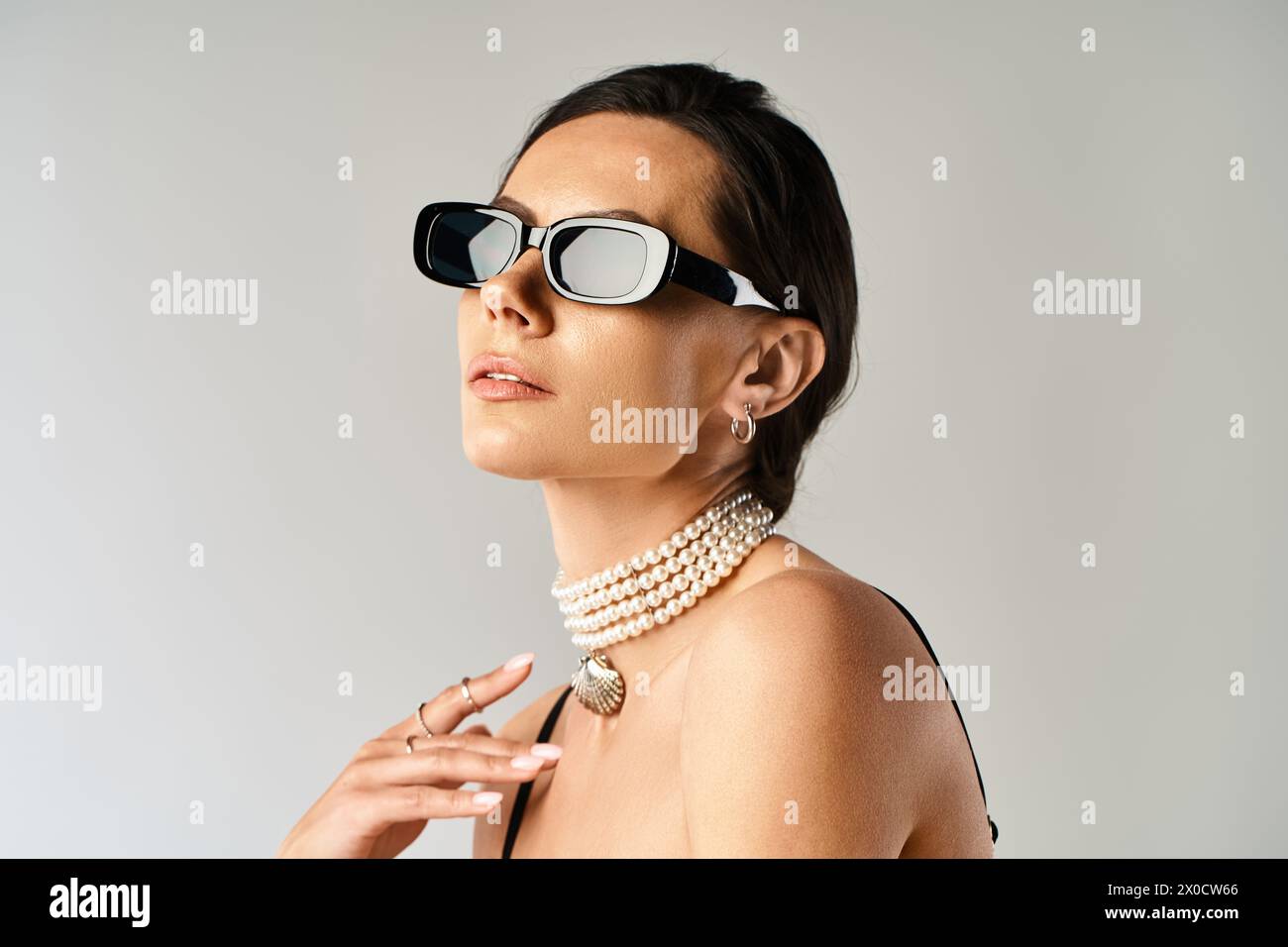 Eine stilvolle Frau mit Sonnenbrille und Perlenkette posiert in einem Studio-Setting vor grauem Hintergrund. Stockfoto