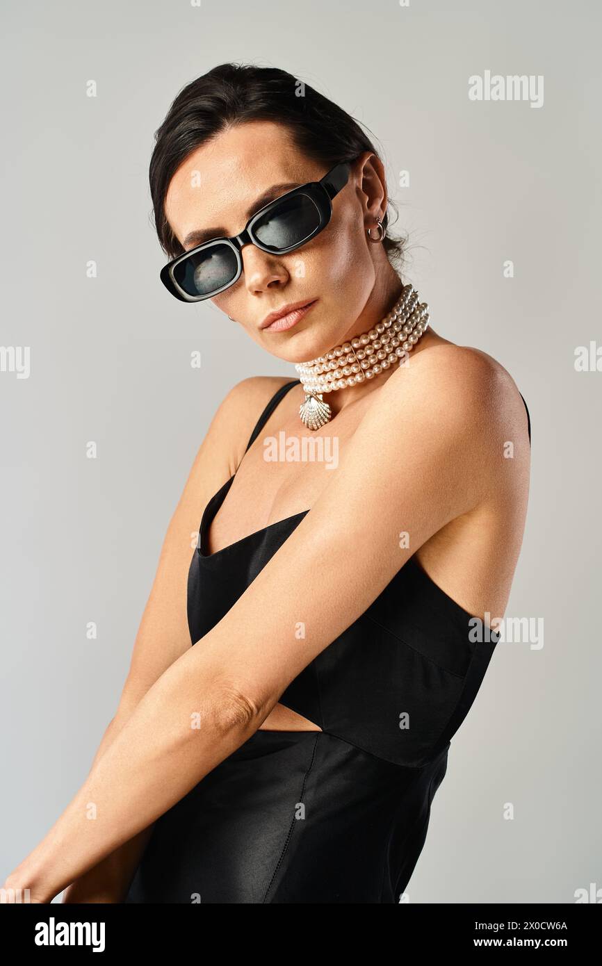 Eine stilvolle Frau verströmt Selbstvertrauen in einem schwarzen Kleid, das mit einer schicken Sonnenbrille ausgestattet ist und in einem Studio vor grauem Hintergrund steht. Stockfoto