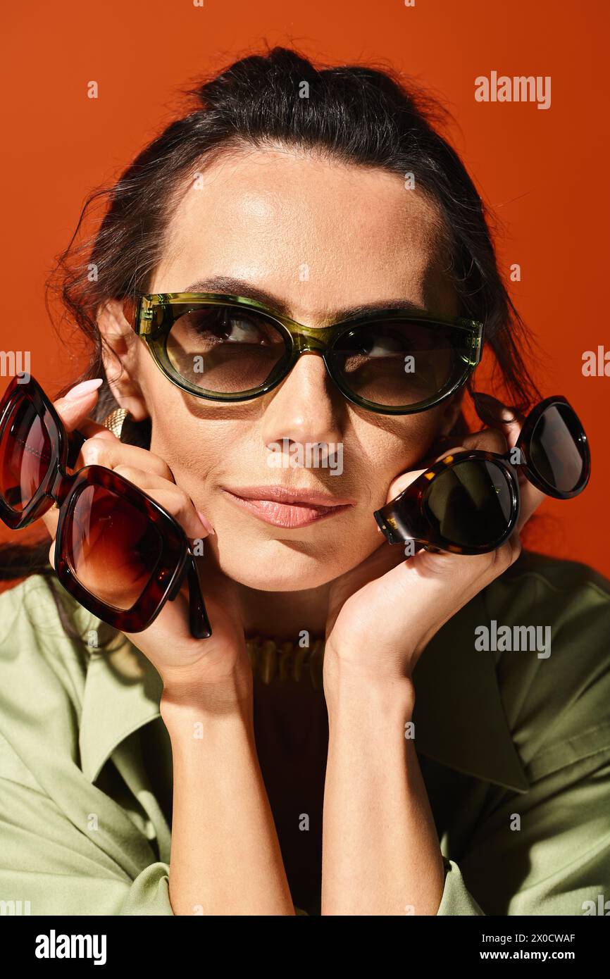 Eine stilvolle Frau, die eine Sonnenbrille trägt und mit den Händen im Gesicht Selbstvertrauen und Sommeratmosphäre auf einem orangefarbenen Studiohintergrund ausstrahlt. Stockfoto