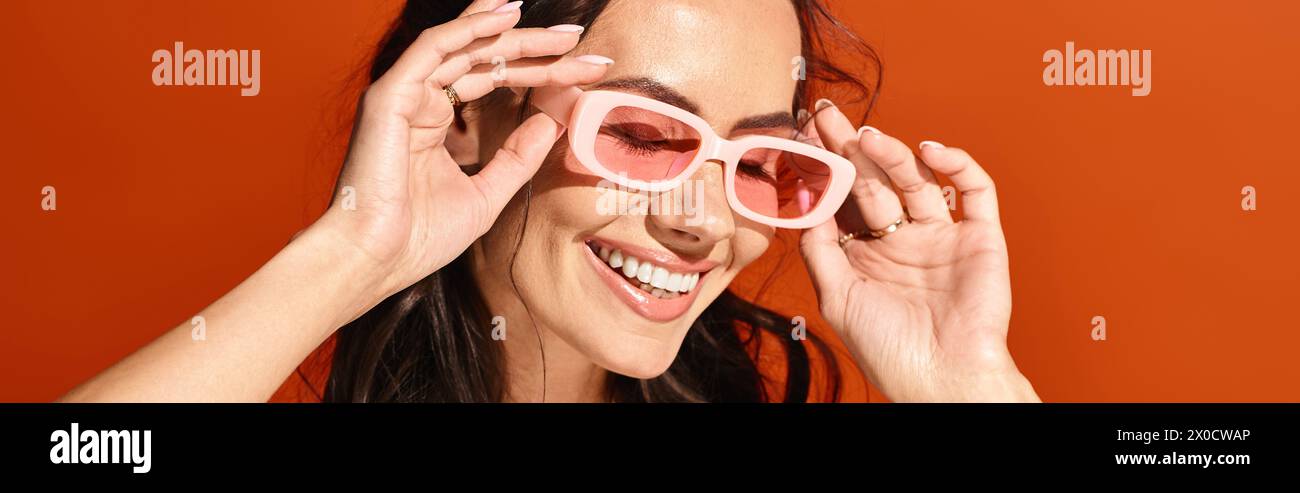 Eine hübsche Frau strahlt, während sie eine rosafarbene Sonnenbrille in Herzform vor einem orangefarbenen Studio-Hintergrund trägt. Stockfoto