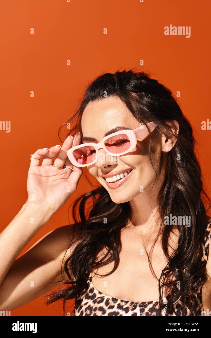 Eine stilvolle Frau strahlt Vertrauen aus in einer rosa Sonnenbrille und einem Oberteil mit Leopardenmuster auf orangefarbenem Hintergrund. Stockfoto