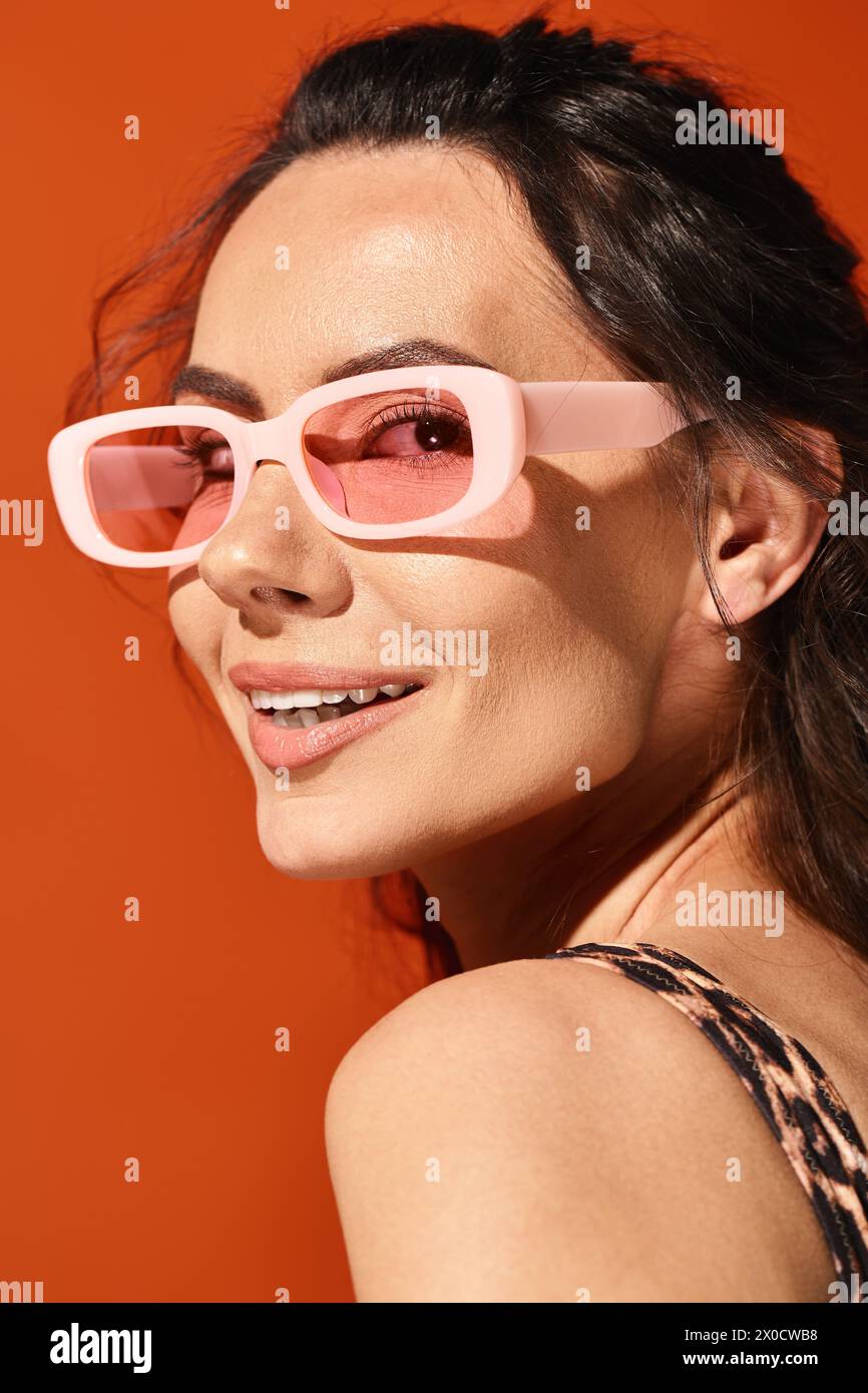 Eine hübsche Frau lächelt, während sie in einem Studio eine pinkfarbene Sonnenbrille vor einem orangen Hintergrund trägt und die Sommermode zeigt. Stockfoto