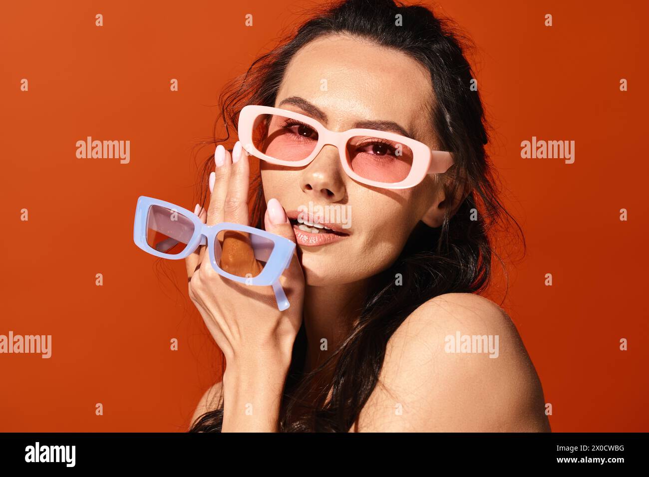 Eine stilvolle Frau in rosafarbenen Gläsern hält eine Sonnenbrille lässig vor einem leuchtenden orangefarbenen Hintergrund. Stockfoto