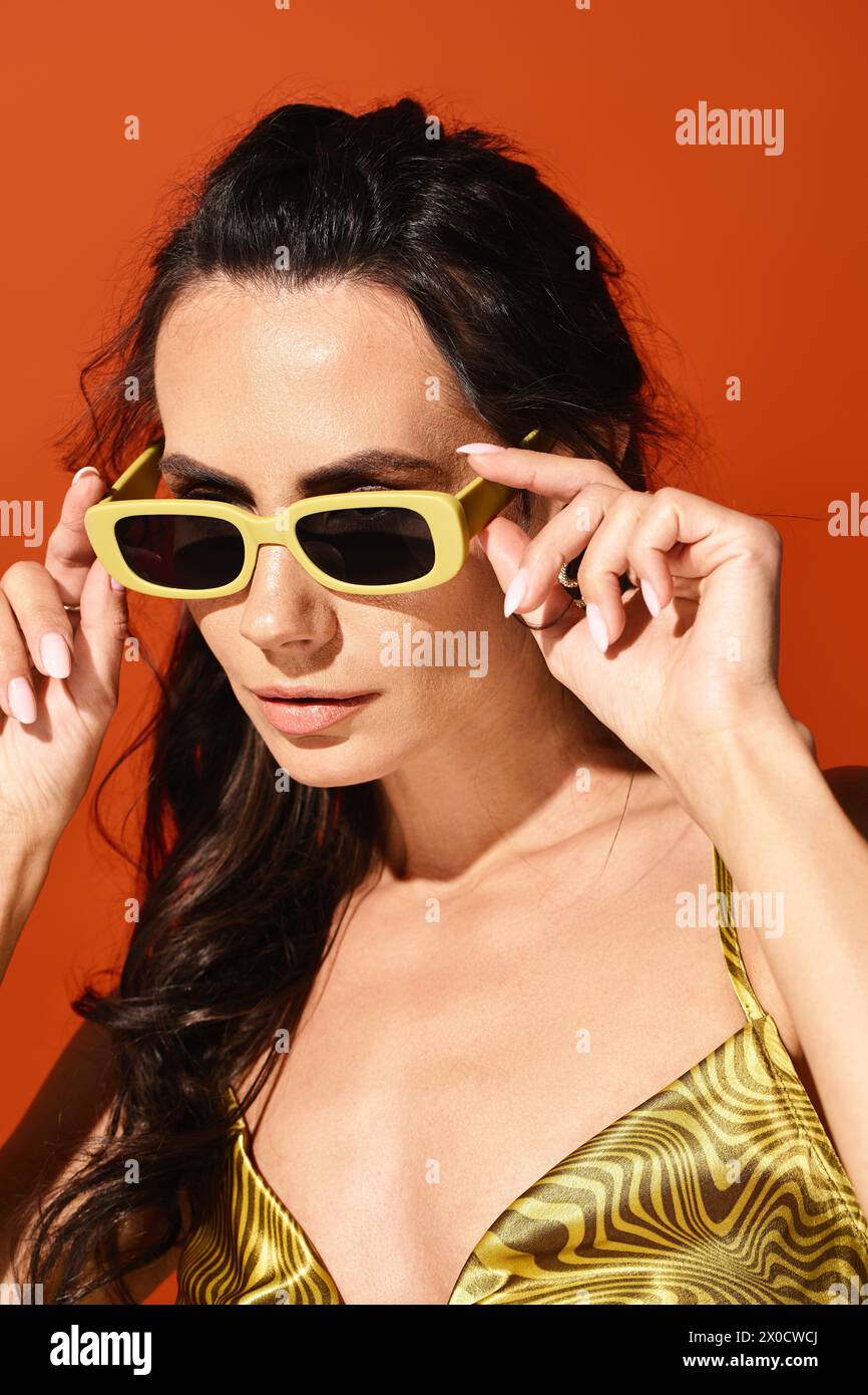 Eine stilvolle Frau mit strahlendem Lächeln trägt ein leuchtendes gelbes Oberteil und eine trendige Sonnenbrille vor einem auffälligen orangefarbenen Hintergrund. Stockfoto