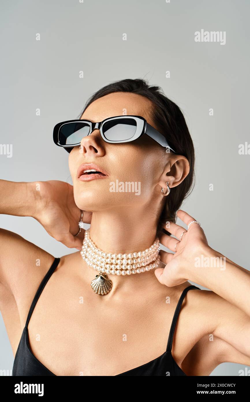 Eine stilvolle Frau strahlt Vertrauen in eine Sonnenbrille und eine Halskette vor grauem Hintergrund aus. Stockfoto