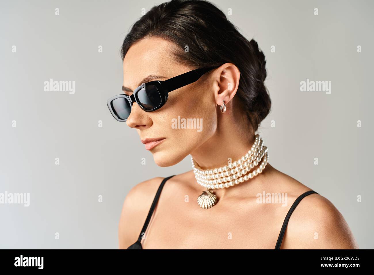 Eine modische Frau mit stilvoller Sonnenbrille und schicker Halskette posiert in einem Studio vor grauem Hintergrund. Stockfoto