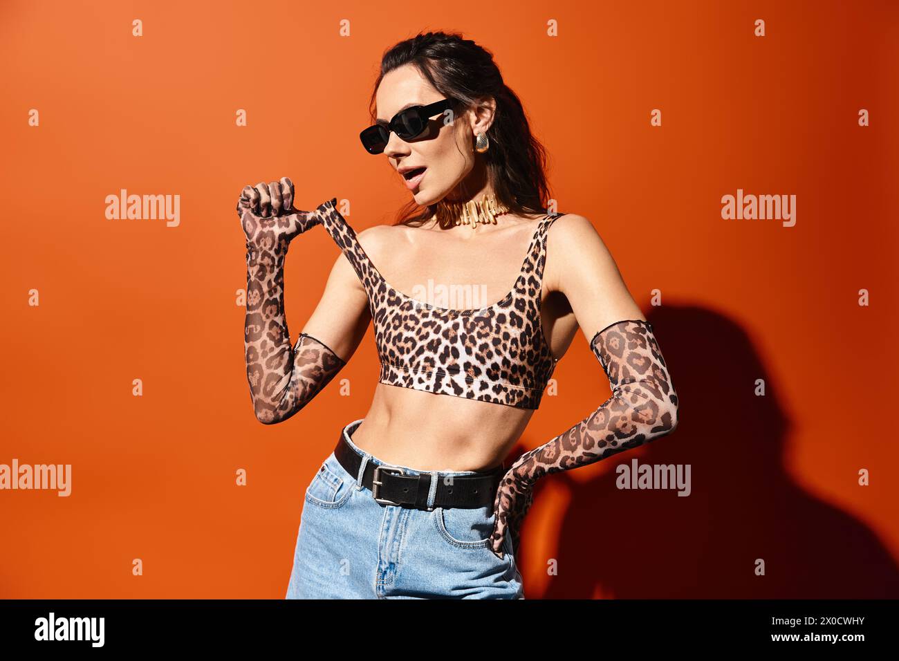 Eine modische Frau trägt selbstbewusst ein Oberteil mit Leopardenmuster und eine Jeans mit Sonnenbrille vor orangefarbenem Studiohintergrund. Stockfoto