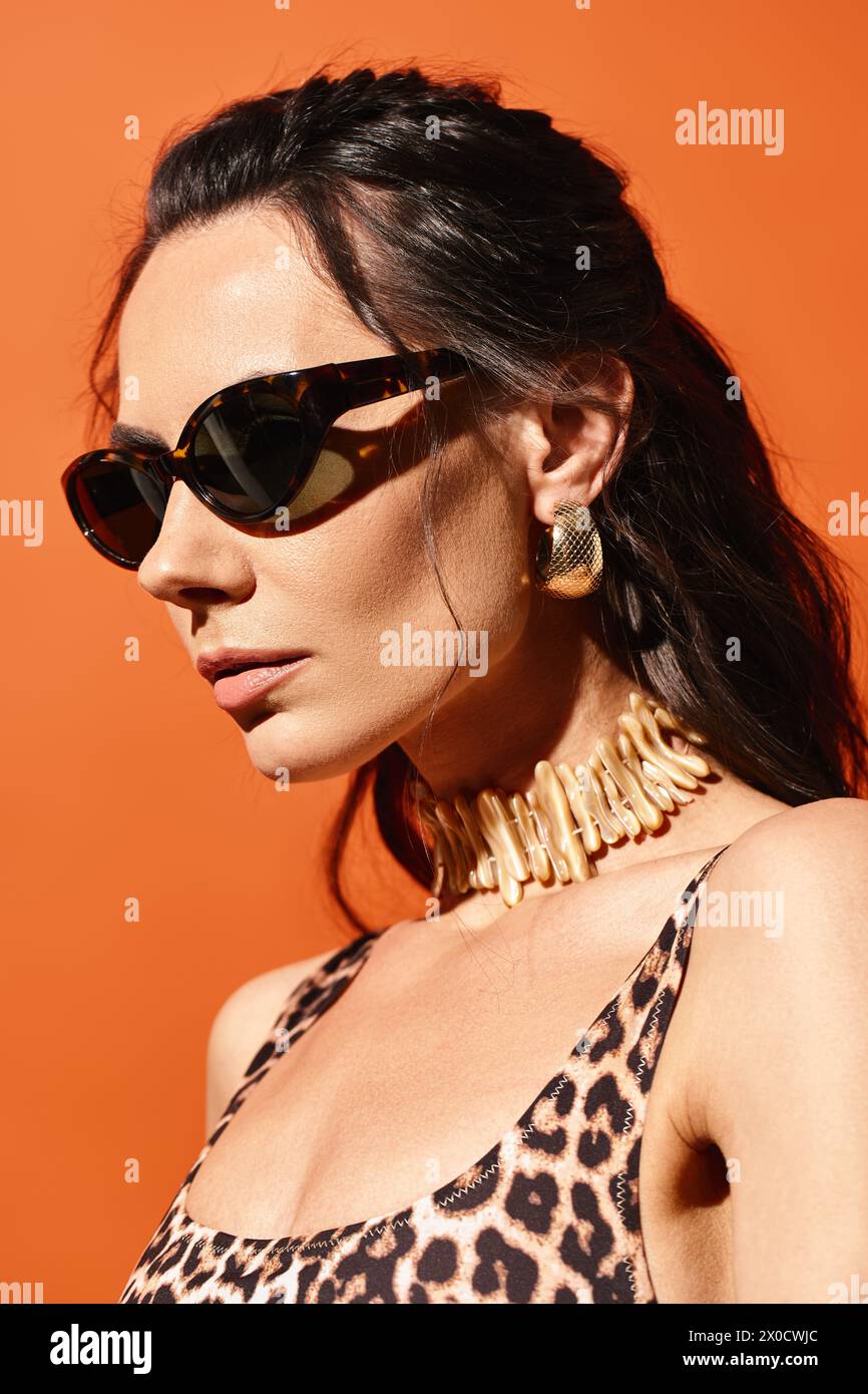 Eine stilvolle Frau mit Sonnenbrille posiert in einem Oberteil mit Leopardenmuster vor orangem Studiohintergrund und strahlt im Sommer Mode-Flair aus. Stockfoto