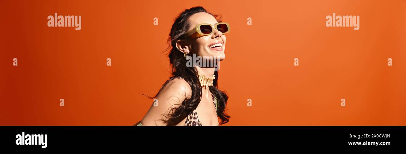 Eine stilvolle Frau in einem Kleid mit Leopardenmuster und einer Sonnenbrille posiert selbstbewusst in einem Studio vor einem orangefarbenen Hintergrund. Stockfoto