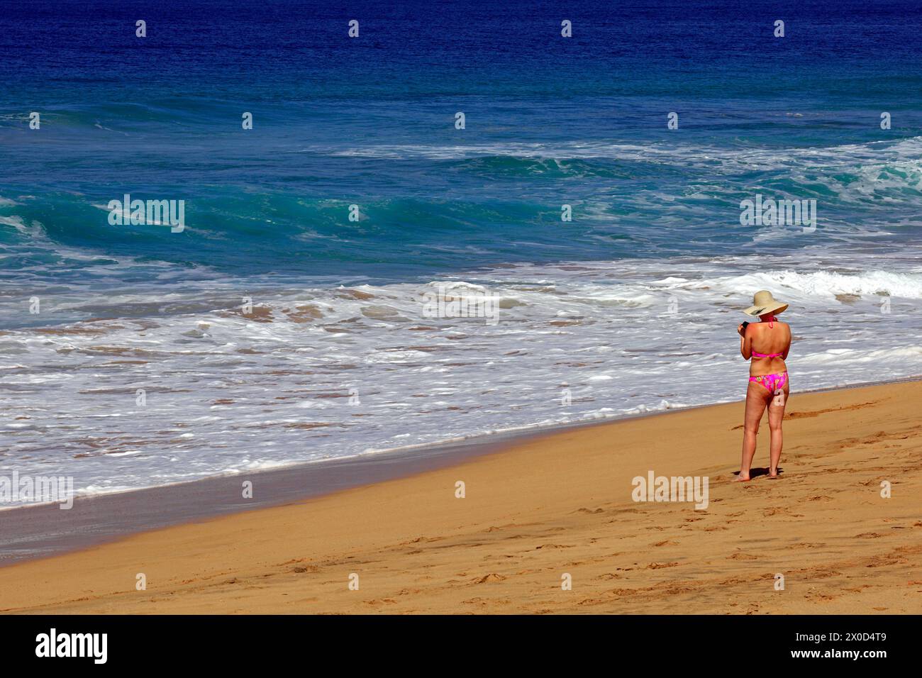 Frau in Bikini und Sonnenhut mit Handy am Strand, El Cotillo, Fuerteventura. Vom Februar 2024 Stockfoto