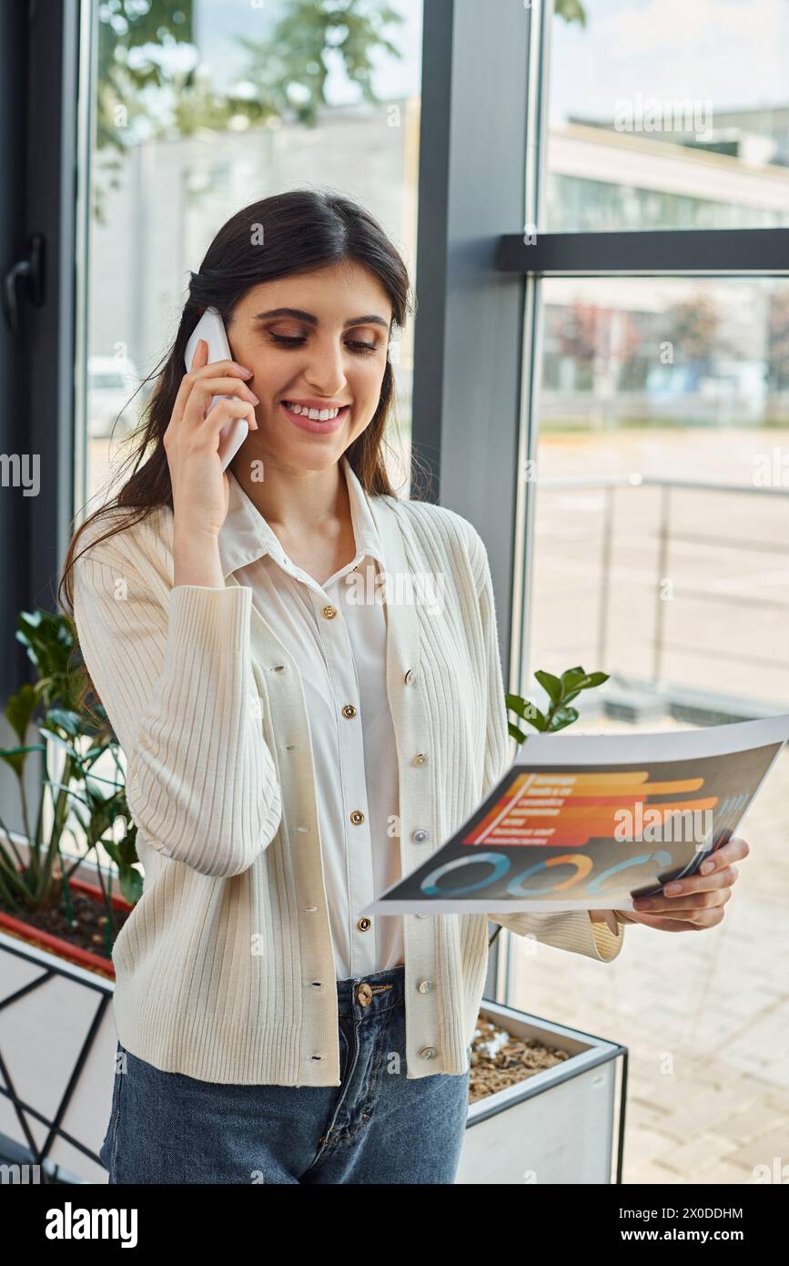 Eine Geschäftsfrau, die in einem modernen Büroumfeld mit einem Mobiltelefon telefoniert, während sie Karten hält. Stockfoto