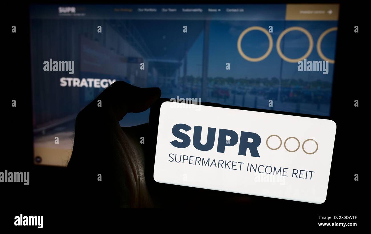 Person, die ein Mobiltelefon mit dem Logo der britischen Immobiliengesellschaft Supermarket Income REIT plc vor der Webseite hält. Schwerpunkt auf dem Display des Telefons. Stockfoto